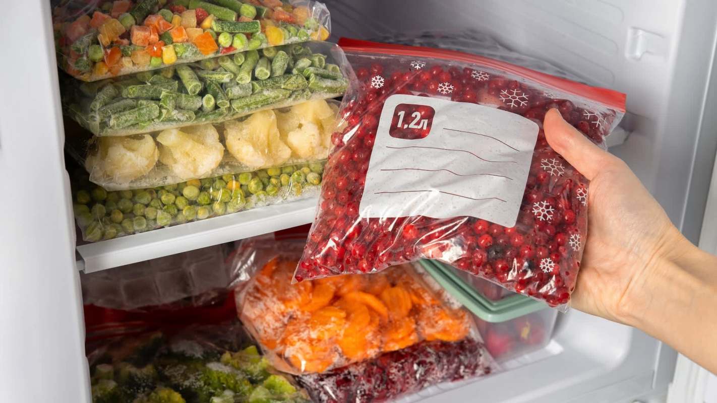 A hand reaches for a bag of red berries in a freezer filled with assorted vegetables and fruits.
