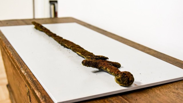 An ancient, rusted sword displayed on a white surface with a wooden table underneath.