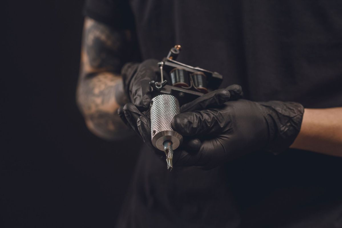 A person wearing gloves holds a tattoo machine and a tattoo needle against a dark background.