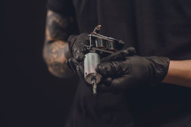 A person wearing gloves holds a tattoo machine and a tattoo needle against a dark background.