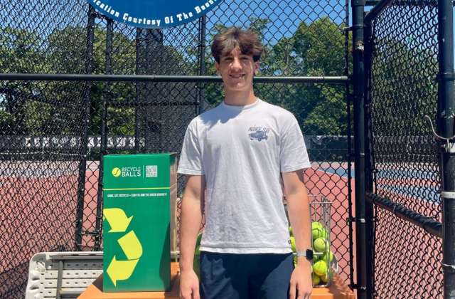 A young man stands beside a green recycling bin for used tennis balls at a tennis court.