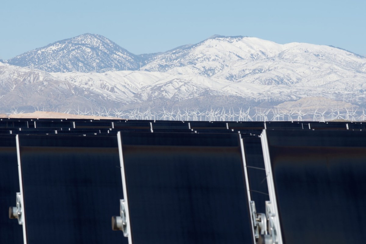 A row of solar panels in front of wind turbines and snow-capped mountains.