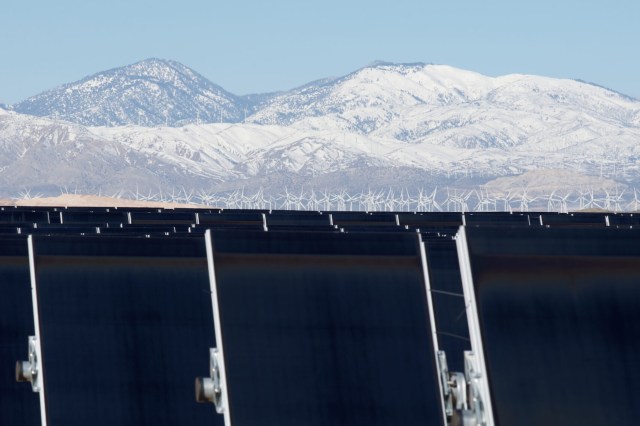 A row of solar panels in front of wind turbines and snow-capped mountains.