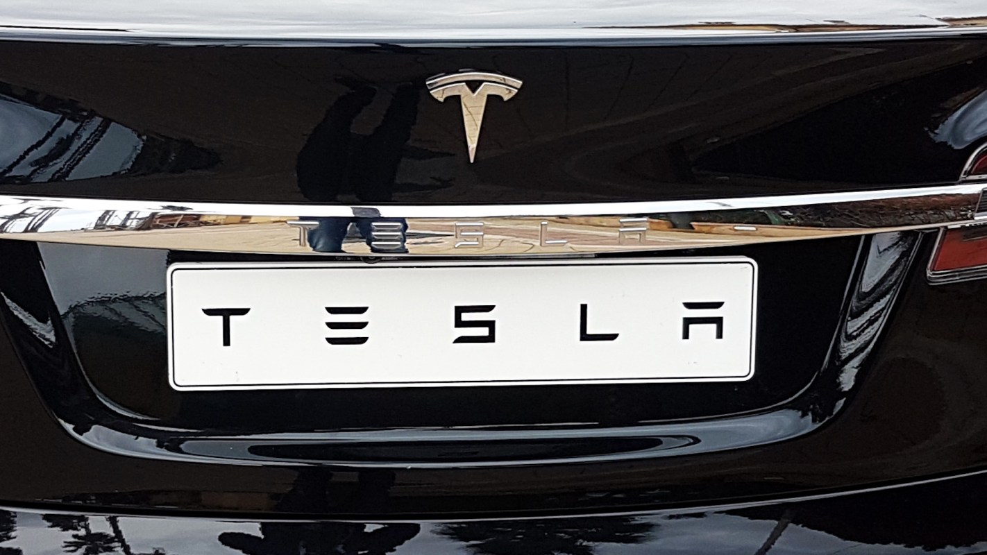 A close-up of the back of a black Tesla car showing the logo and license plate.