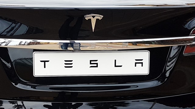 A close-up of the back of a black Tesla car showing the logo and license plate.