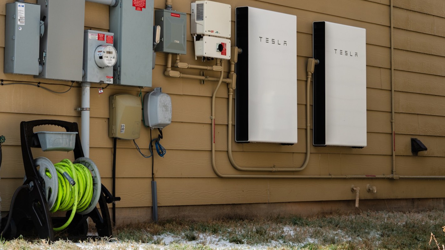 Two Tesla battery units mounted on an exterior wall next to electrical panels and a garden hose reel.