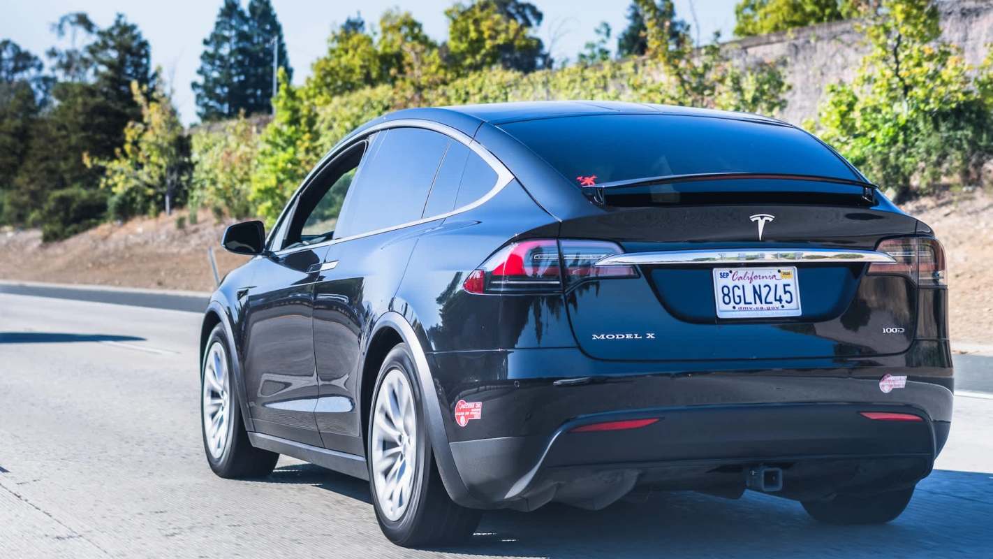 A black Tesla Model X driving on a highway with greenery in the background.