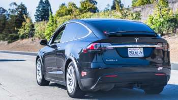 A black Tesla Model X driving on a highway with greenery in the background.