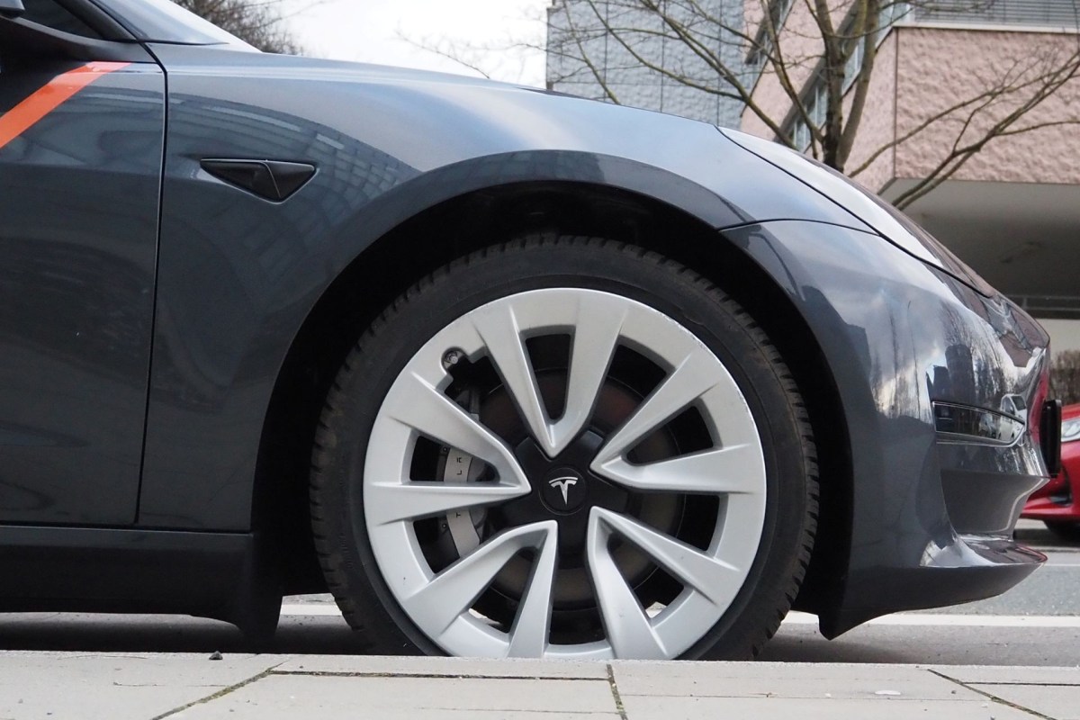 A close-up view of a Tesla car's wheel and side, showcasing the gray paint and distinctive alloy rim design.