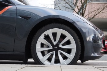 A close-up view of a Tesla car's wheel and side, showcasing the gray paint and distinctive alloy rim design.