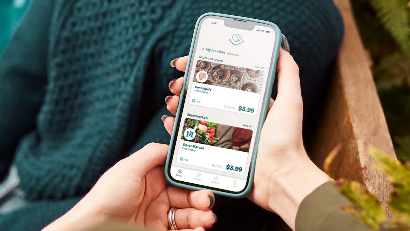 A person holds a smartphone displaying a Too Good To Go food delivery app with nearby store options and prices.