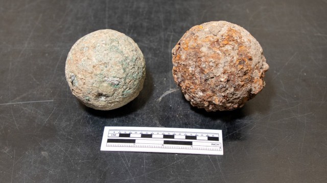 Two historical spherical objects, one rusty and one oxidized, placed beside a measurement scale on a dark surface.
