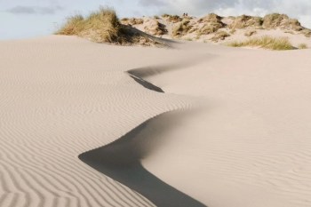 Gentle dunes of sand undulate beneath a blue sky with sparse clouds, creating a textured landscape.