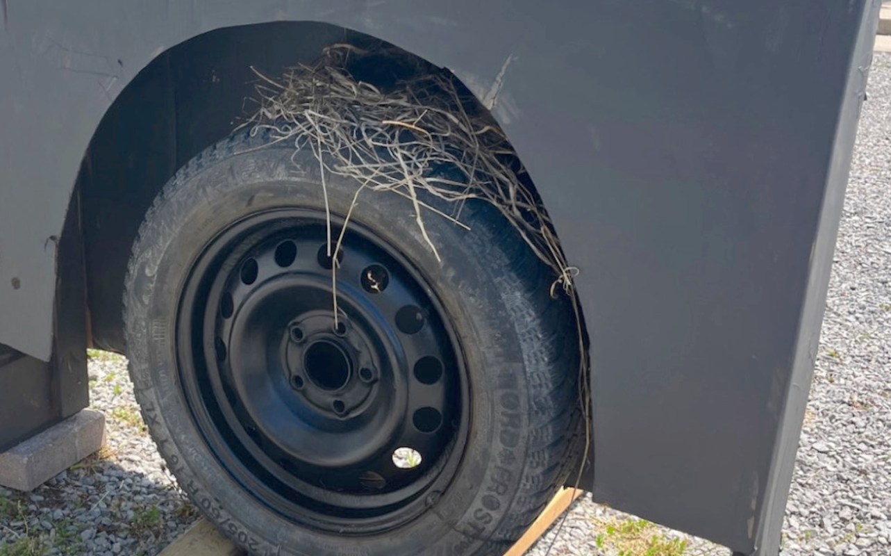 A black tire with dried twigs and grass woven into it sits beside a gray vehicle base on gravel.