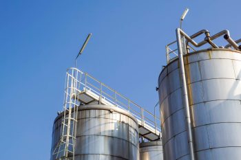 Two large industrial silos with a metal ladder and piping against a clear blue sky.