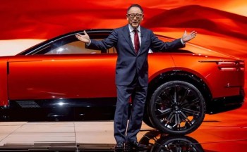 A presenter in a suit stands in front of a sleek red car, gesturing enthusiastically against a vibrant backdrop.