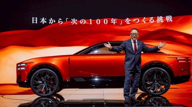 A presenter in a suit stands in front of a sleek red car, gesturing enthusiastically against a vibrant backdrop.