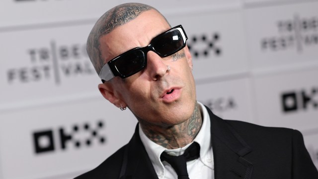 Travis Barker poses in a black suit and sunglasses at a promotional event.