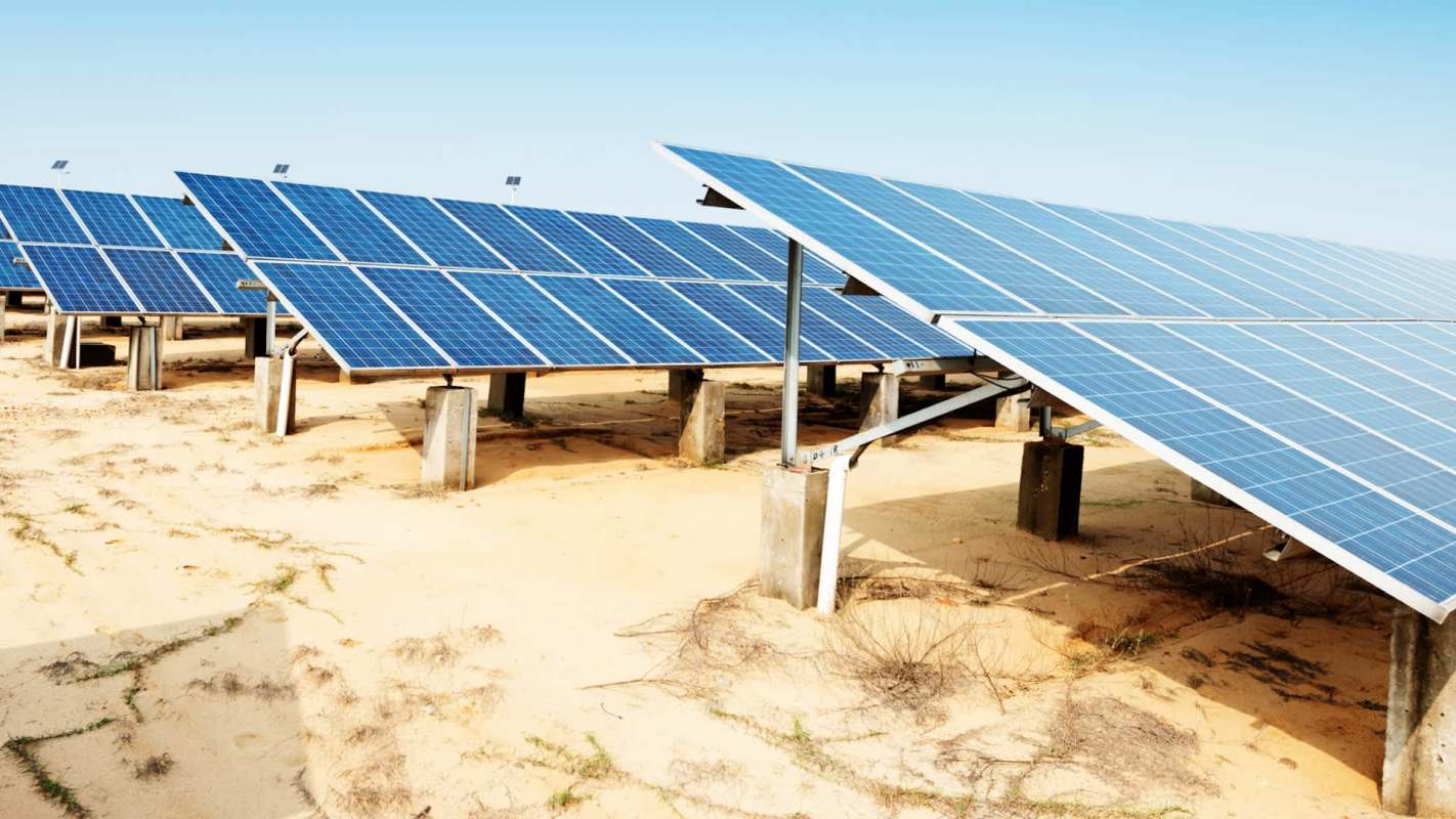 Solar panels arranged in rows on a sandy area under a clear blue sky.