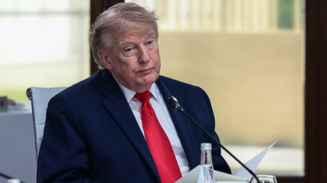 Donald Trump sits at a table in a suit and red tie.