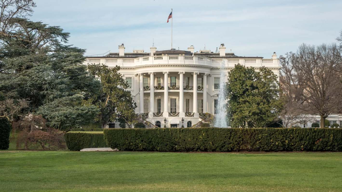 The White House is seen from the garden, featuring lush greenery and a fountain in front.