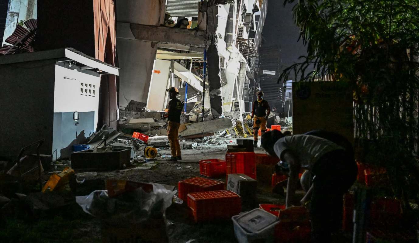 Rescue workers assess damage at a partially collapsed building amidst debris and scattered supplies at night.
