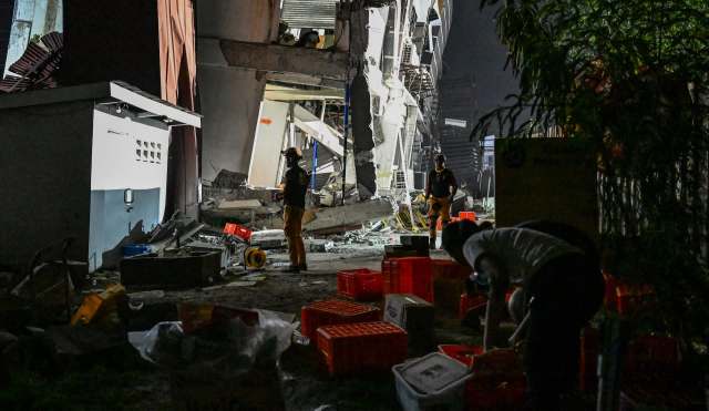 Rescue workers assess damage at a partially collapsed building amidst debris and scattered supplies at night.