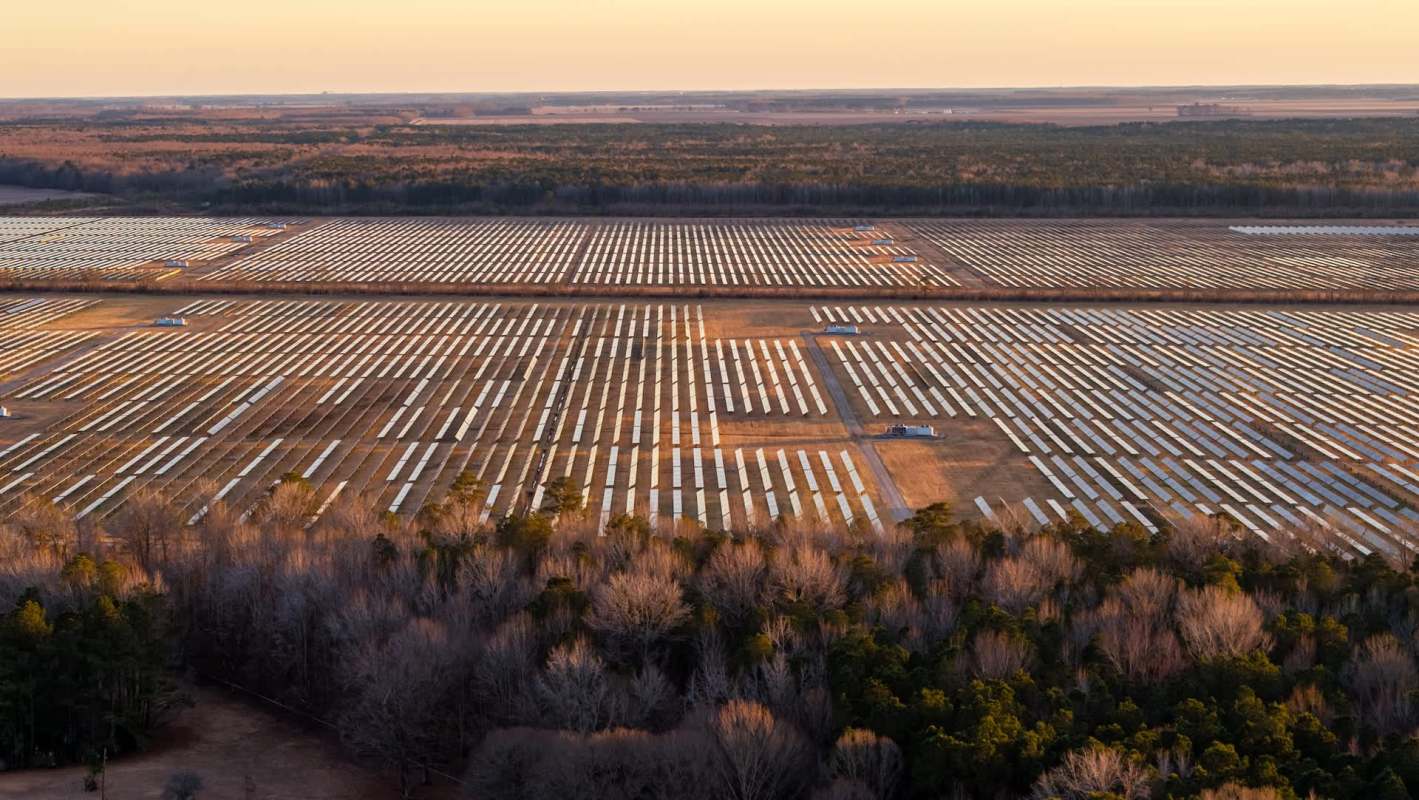 Aerial view of a vast solar farm with numerous solar panels set against a backdrop of trees and fields.