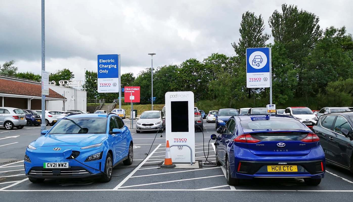 Two electric vehicles at charging stations in a Tesco parking lot.