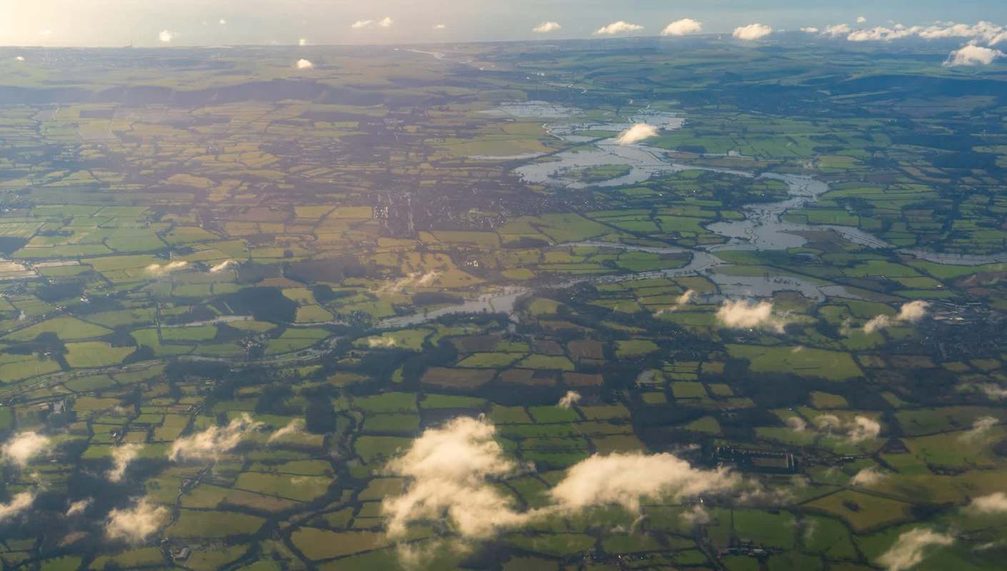 Aerial view of green fields, rivers, and patches of clouds under a clear sky.