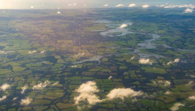 Aerial view of green fields, rivers, and patches of clouds under a clear sky.