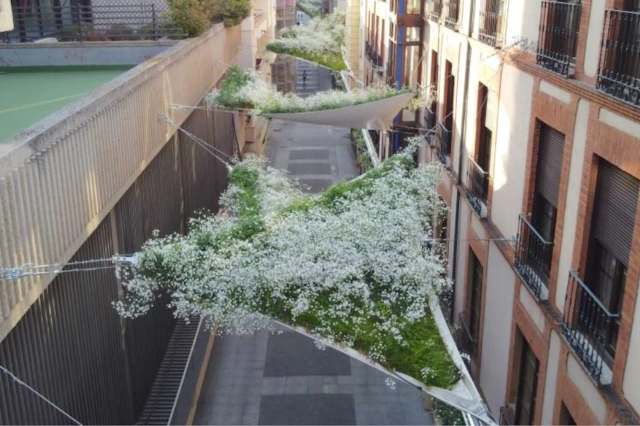 A narrow urban street adorned with suspended greenery and white flowers creating a unique overhead installation.
