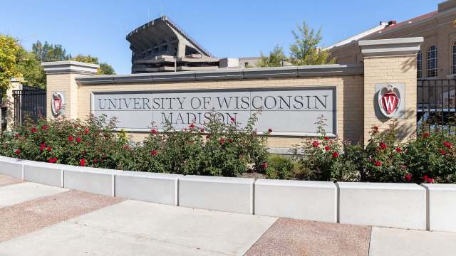 A sign for the University of Wisconsin-Madison surrounded by blooming rose bushes and a stadium in the background.