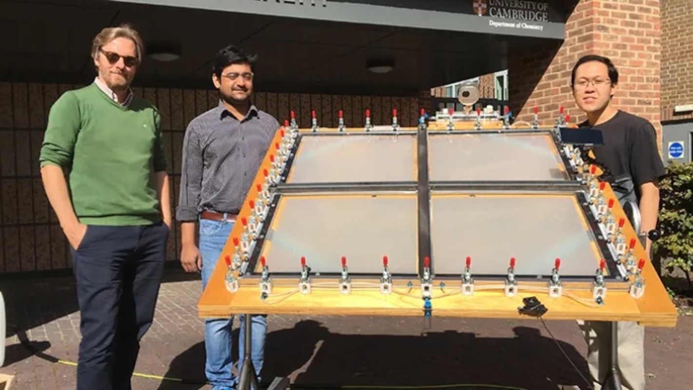 Three men stand beside a large experimental apparatus with multiple valves outside the University of Cambridge building.