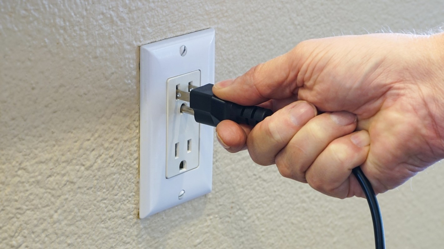 A hand is plugging a black electrical cord into a white wall outlet.