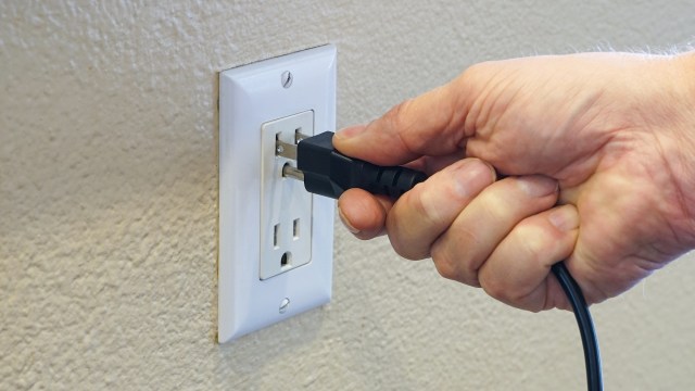 A hand is plugging a black electrical cord into a white wall outlet.