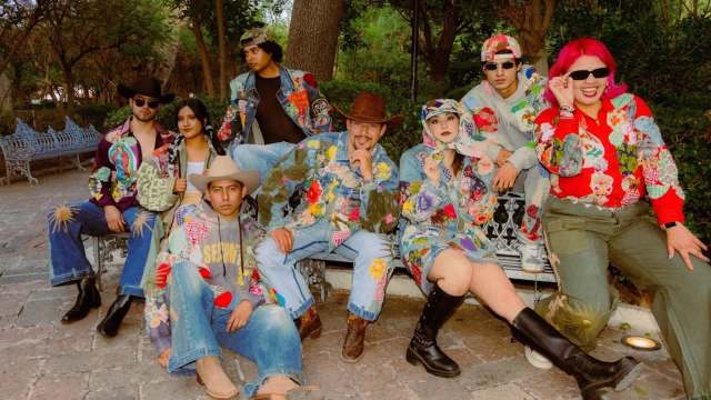 A group of young adults poses together in colorful, embellished denim outfits in a lush park setting.