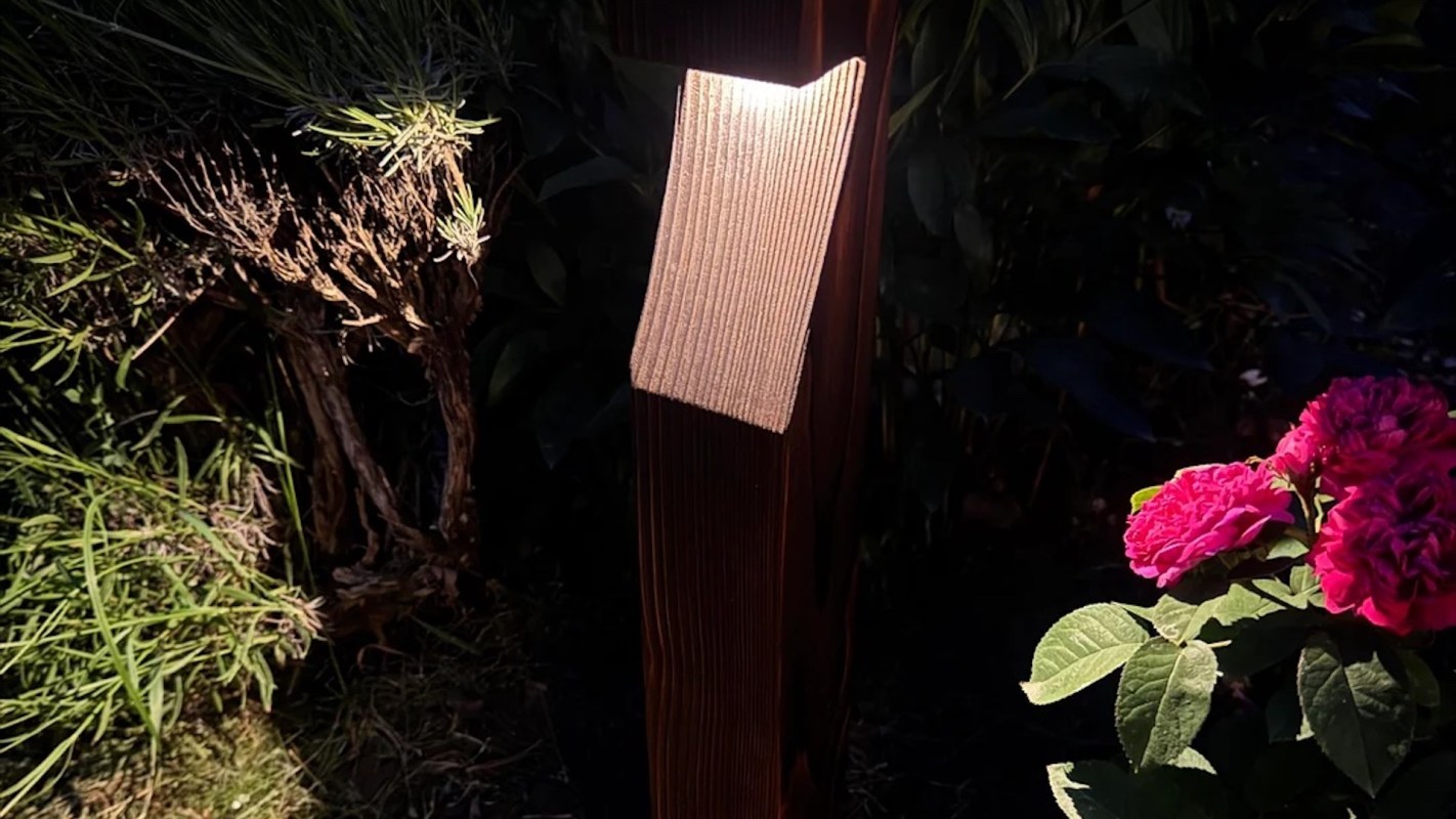 A wooden post with a soft light at the top, surrounded by greenery and pink flowers in a dark garden setting.