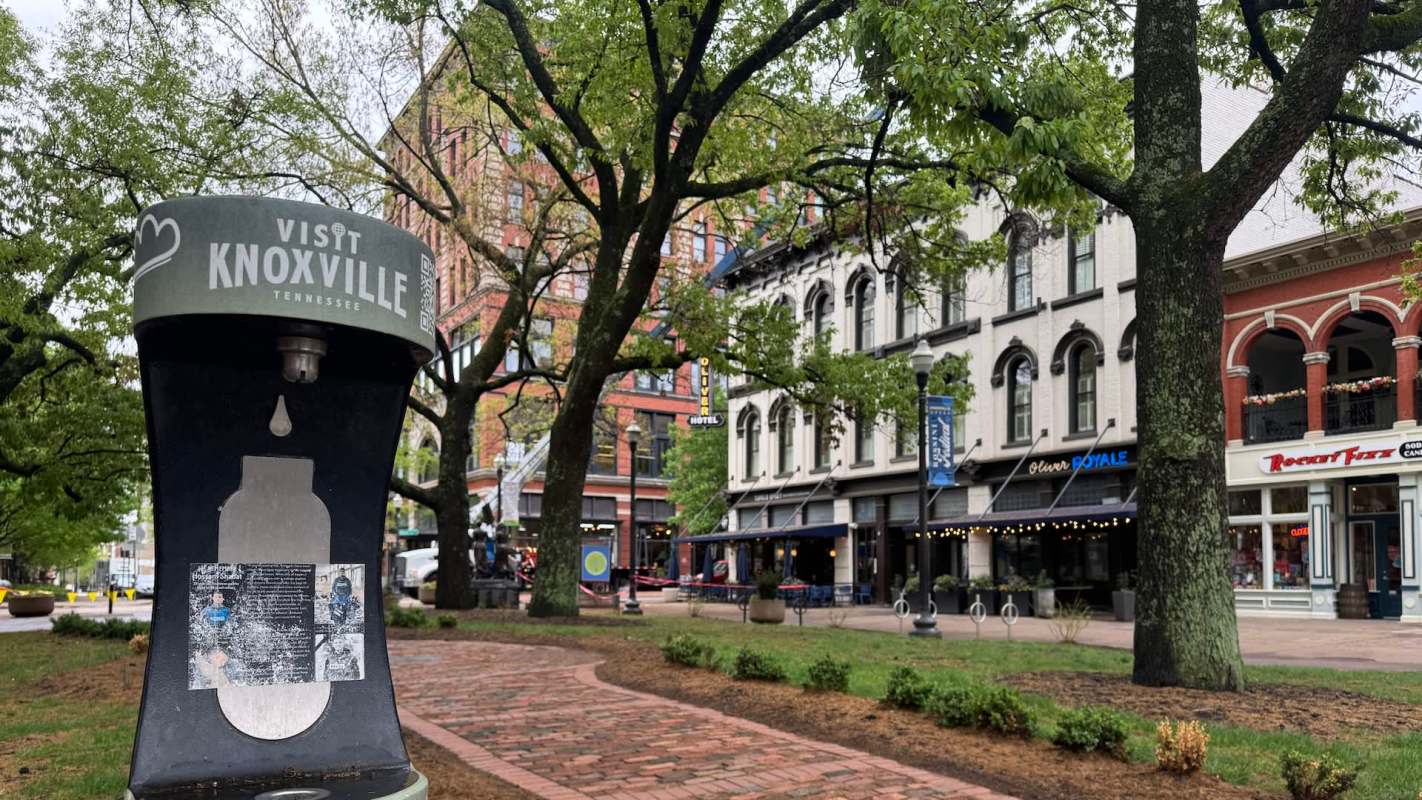 A green informational sign reads "Visit Knoxville" in a city park with trees and historic buildings in the background.