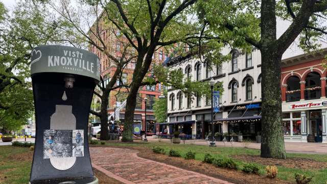 A green informational sign reads "Visit Knoxville" in a city park with trees and historic buildings in the background.