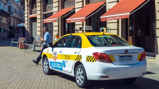 A man walks past an electric taxi parked in a sunlit, vibrant street lined with shops.