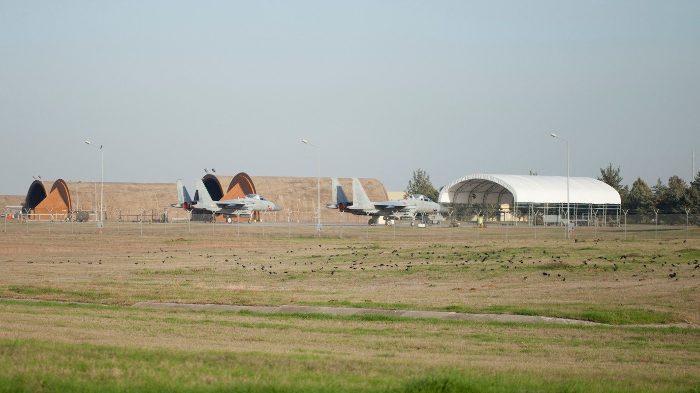 Two military aircraft sit on a runway beside hangars, with a grassy field and trees in the background.
