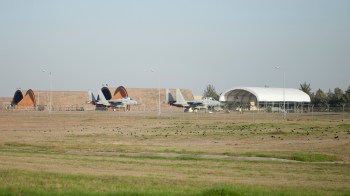 Two military aircraft sit on a runway beside hangars, with a grassy field and trees in the background.