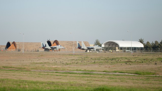 Two military aircraft sit on a runway beside hangars.