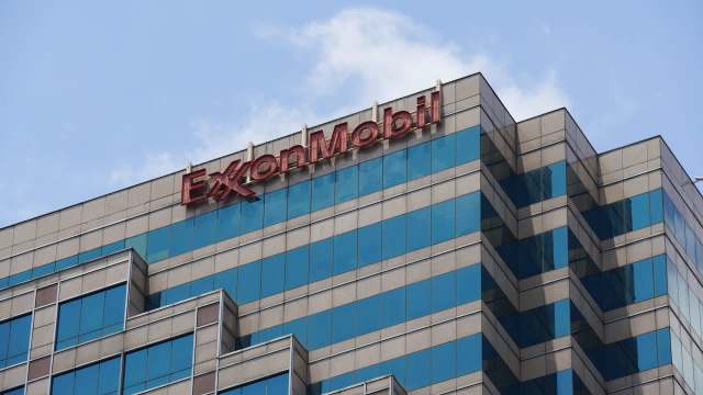A close-up view of the ExxonMobil sign on a modern glass office building against a clear blue sky.