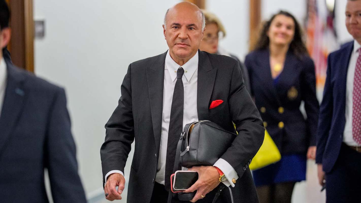 Kevin O'Leary carries a bag and phone while walking through a corridor with others in the background.