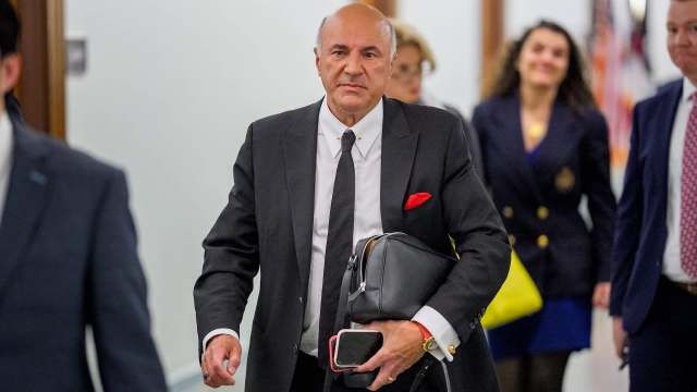 Kevin O'Leary carries a bag and phone while walking through a corridor with others in the background.