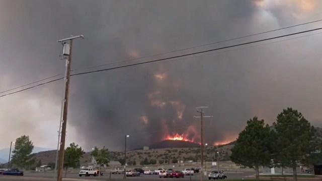 A wildfire burns brightly on a hillside under a dark, smoky sky, with nearby vehicles parked below.