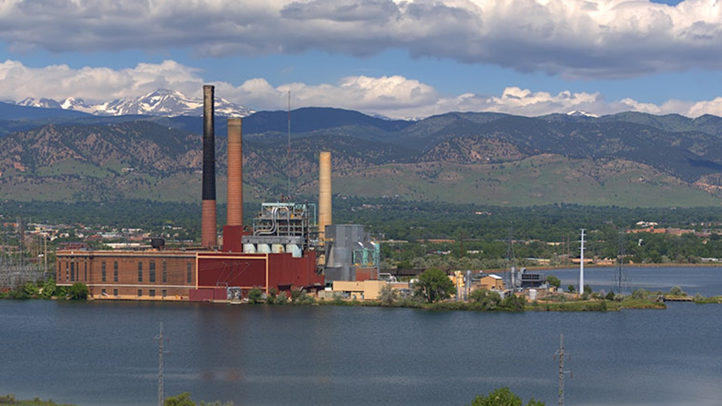 A power plant with tall smokestacks near a calm lake and mountains in the background under a partly cloudy sky.