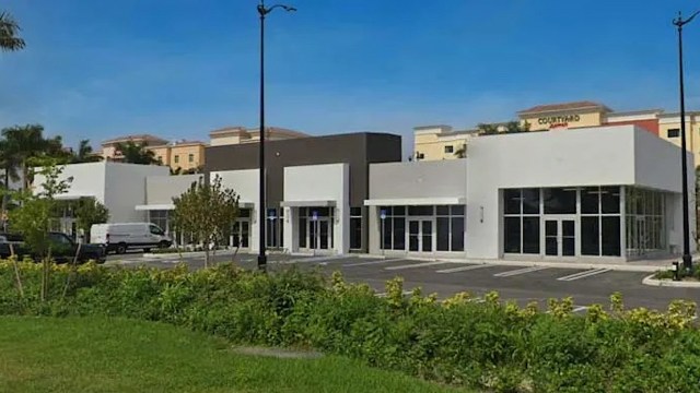 A row of modern commercial buildings with large windows and empty parking spaces under a clear blue sky.
